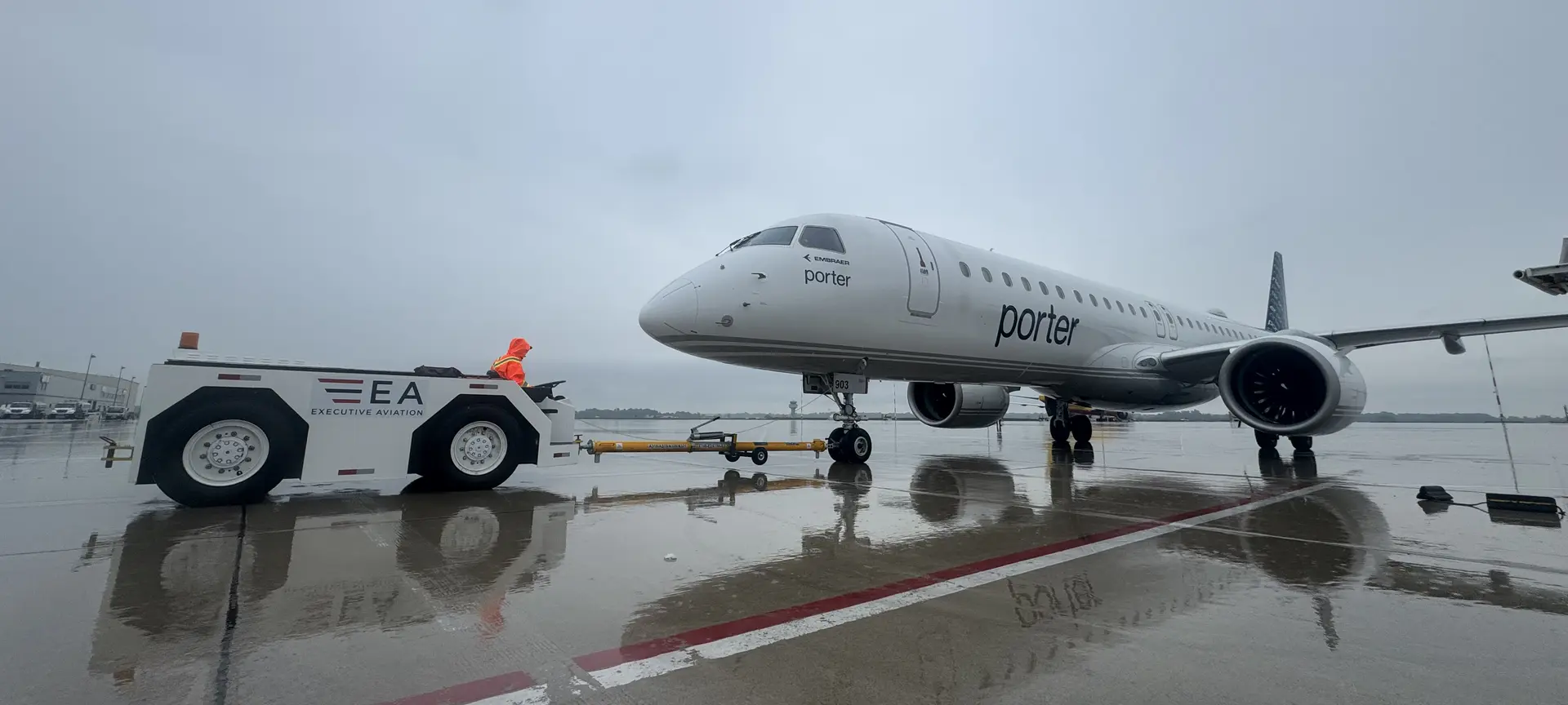 An Enedym-converted electric pushback tractor towing an Embraer E195 across a wet apron